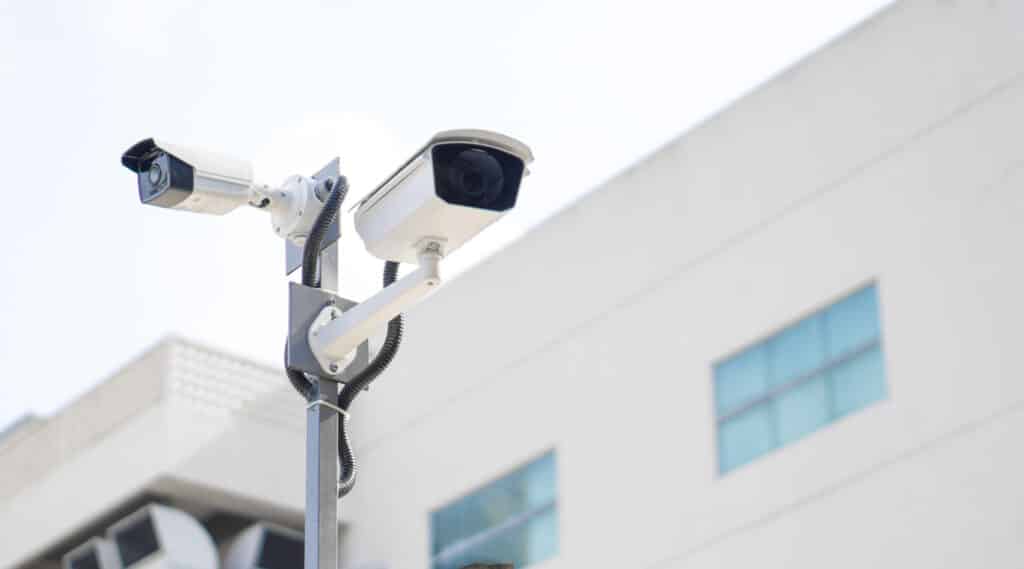 Deux caméras de surveillance blanches montées sur un poteau gris devant un bâtiment blanc, câbles noirs visibles.