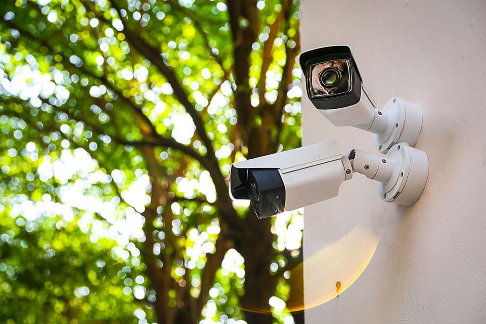 Caméras de surveillance extérieures : sécurité et arbres Deux caméras de surveillance extérieures blanches fixées sur un mur clair, surveillant un fond d'arbres verts flous avec un fort bokeh lumineux.