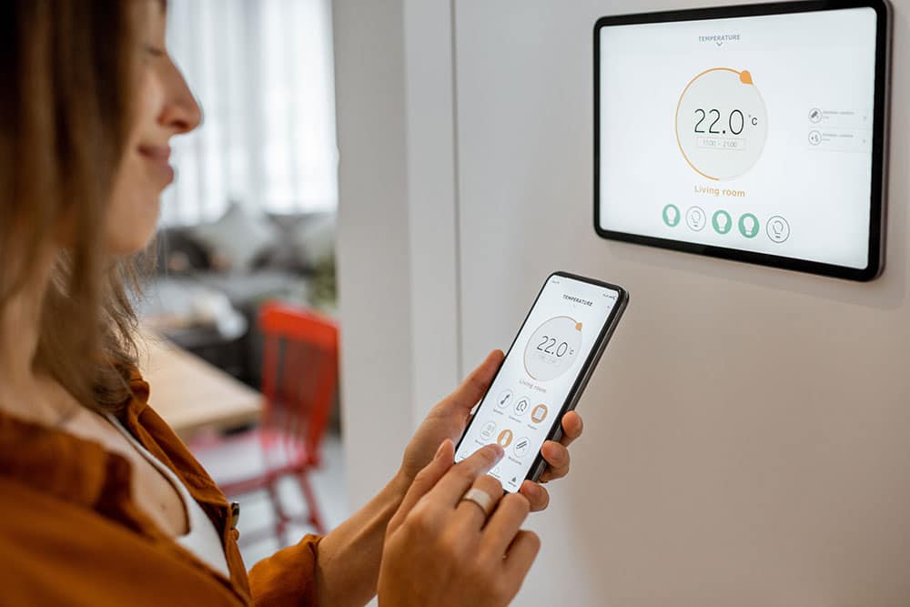Contrôle Domotique : Thermostat Connecté et Smartphone Femme utilisant un smartphone pour contrôler un thermostat mural affichant 22.0°C pour le salon (Living room), en lien avec la domotique.