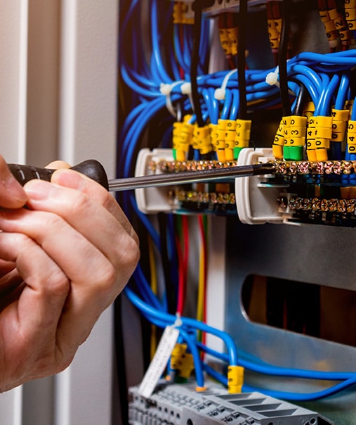 Électricien vérifie le câblage du tableau électrique Mains d'électricien serrant une vis sur un bornier avec un tournevis dans un panneau de contrôle rempli de fils bleus et d'étiquettes jaunes numérotées (0, 1, 2, 3, 4, 5).
