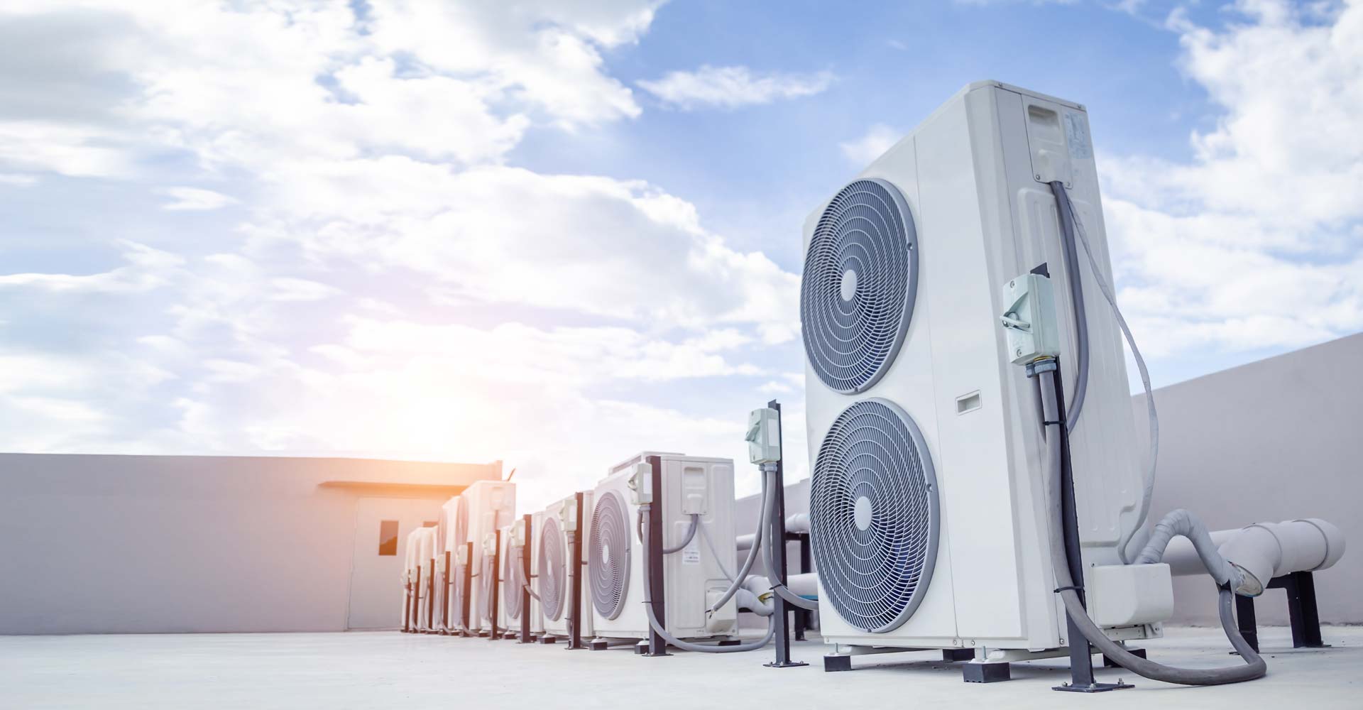 Plusieurs unités extérieures de climatisation, ou thermopompes, alignées sur un toit plat sous un ciel bleu clair et nuageux avec un fort contre-jour.